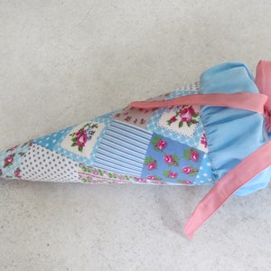Könnte beinhalten: Eine blaue und weiße Patchwork-Stofftasche in Kegelform mit einem rosa Band, das um die Oberseite gebunden ist. Die Tasche ist mit kleinen rosa Blumen verziert.