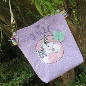 Könnte beinhalten: Eine lavendelfarbene Umhängetasche mit weißen Punkten. Die Tasche hat ein gesticktes Einhorn-Design in einem rosa Kreis, einen hellblauen Schmetterling und den Namen "Juli". Ein kleiner, gestreifter Schlüsselanhänger ist befestigt.
