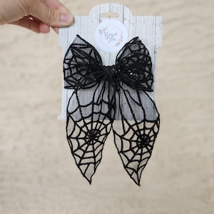May include: Black spiderweb bow with a white background. The bow is made of mesh fabric and has a glittery finish. The bow is attached to a white card with the text "Blessed Little" in a pink circle.