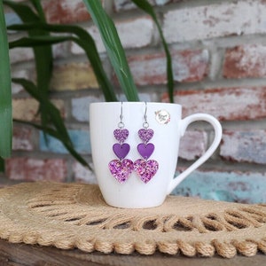 May include: Purple glitter heart dangle earrings with three hearts on each earring. The hearts are layered with the largest heart at the bottom and the smallest heart at the top. The earrings are hanging from a white mug.
