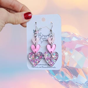 May include: Pink heart-shaped earrings with glitter accents. The earrings are made of layered acrylic and feature a dangling design. The top hearts are a solid light pink, while the bottom hearts are filled with glitter. The earrings are displayed on a white card with the text "Blessed Stitch Studio".