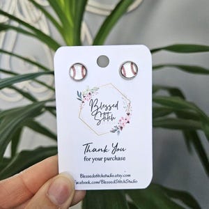 May include: A pair of baseball-themed earrings with white and red baseballs on silver posts. The earrings are displayed on a white card with the text "Blessed Stitch Studio" and "Thank You for your purchase".