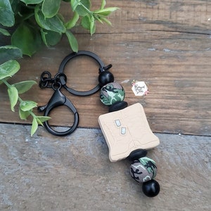 May include: A black keychain with a beige book charm and camouflage beads. The book charm has two small white squares on the front.