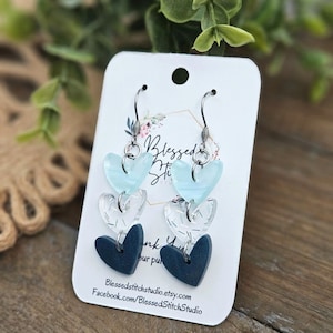 May include: A pair of dangle earrings with three layered hearts. The top heart is light blue, the middle heart is clear with a white pattern, and the bottom heart is dark blue. The earrings are on a white card with the text "Blessed Stitch Studio" and a website address.