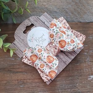 May include: Two pumpkin-themed hair bows with a white background and orange pumpkins. The bows are made with glitter fabric and have a rose gold color. The bows are displayed on a wooden background with a tag that says "Blessed Stitch".