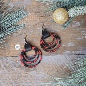 May include: A pair of dangle earrings with three layered circles. The circles are made of red and black plaid fabric and have a black leather accent at the top.