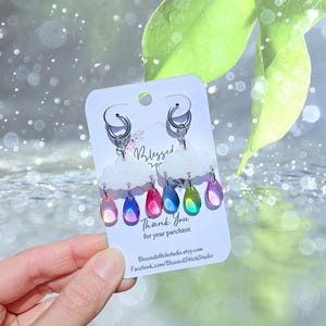 May include: A pair of earrings featuring a cloud design with colorful, iridescent raindrop pendants. The earrings are attached to silver hoops and are displayed on a white card with the text "Blessed" and "Thank you for your purchase."