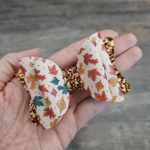 May include: A white bow with a fall leaf print and gold glitter accents. The bow is made of fabric and has a  loop on the back for attaching to hair.