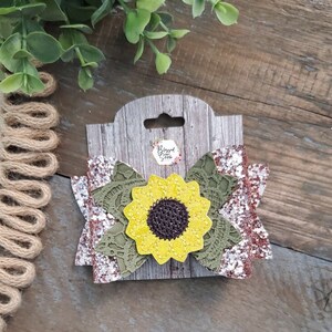 May include: A brown, white, and green glitter bow with a yellow sunflower in the center. The bow is on a wooden background with the text "Blessed Gifts" on a white tag.