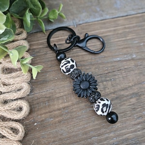 May include: Black beaded keychain with a black flower, leopard print beads, and a black clasp. The keychain is made of various beads and a black metal clasp. The design includes a black flower and leopard print beads.