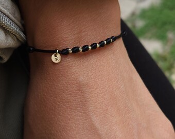May include: Black bracelet with gold-colored beads and a small gold charm with the letter "K". The bracelet is made of a black cord and is worn on the wrist. A simple and elegant piece of jewelry.