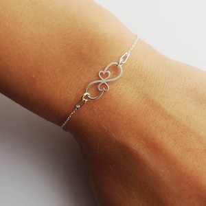 Puede incluir: Una pulsera de plata con un dije de símbolo de infinito hecho de dos corazones entrelazados.