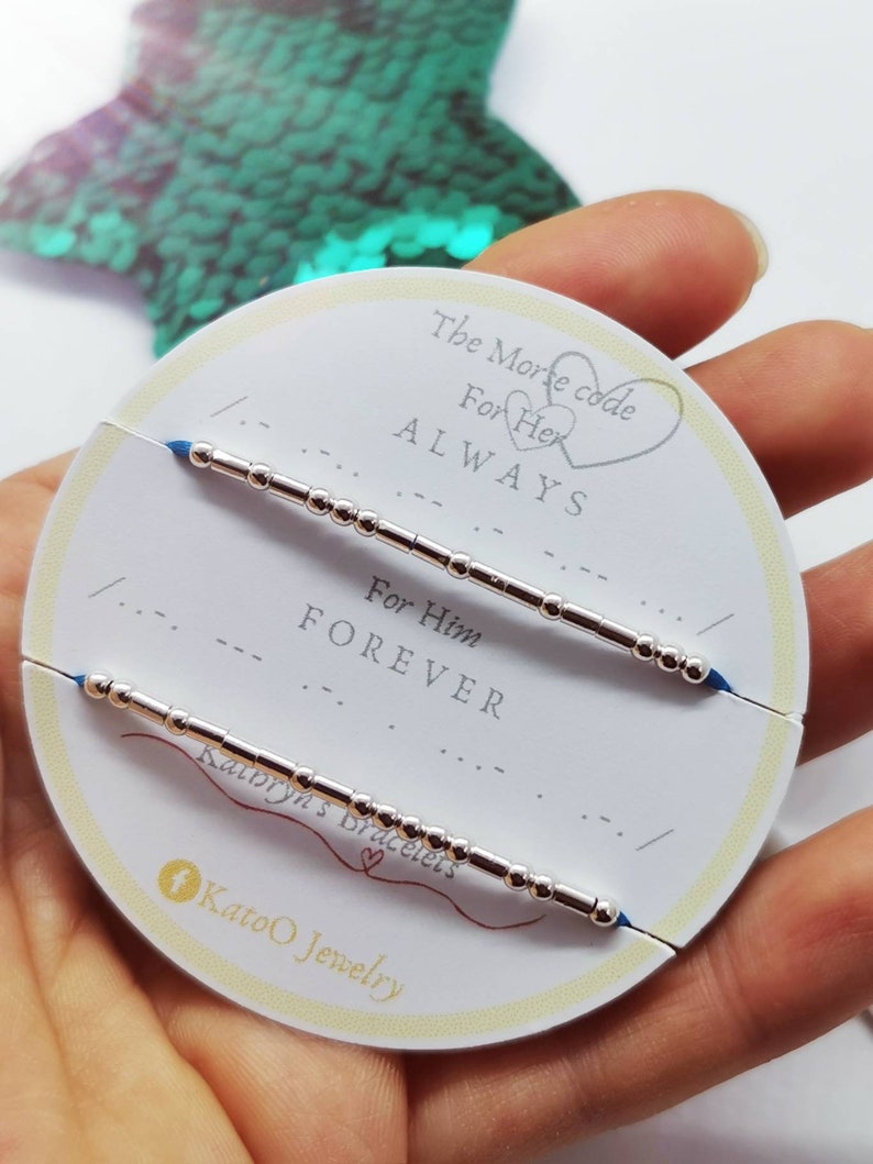 May include: Two silver bracelets with blue string ends. The bracelets are on a white card with the text "The Morse code" and "For Her ALWAYS" and "For Him FOREVER".