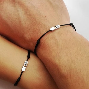May include: Two black string bracelets with silver and gold arrow charms. The bracelets are tied on the wrists of two people.