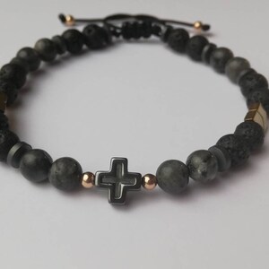 May include: Black beaded bracelet with a gold cross charm. The bracelet is made with lava rock and hematite beads.