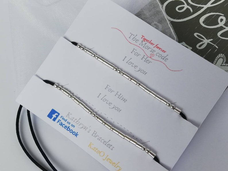 May include: Two black cord bracelets with silver beads spelling out "I love you" in Morse code. The bracelets are displayed on a white card with the text "Together forever. The Morse code. For Her. I love you. For Him. I love you. Kathryn's Bracelets. Find us on Facebook. Katoo Jewelry."