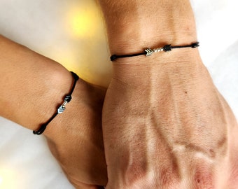 Puede incluir: Dos pulseras de cuerda negra con dijes de flecha plateada. Las pulseras se llevan en las muñecas de dos personas.