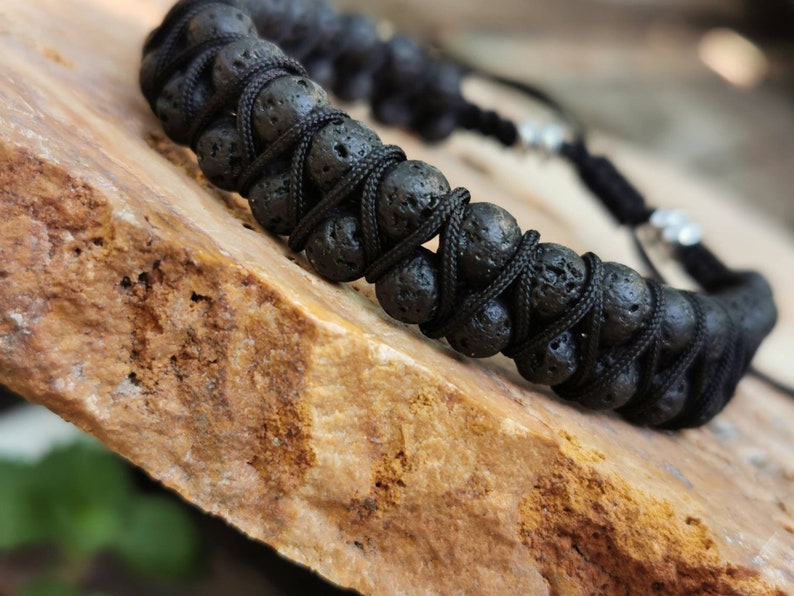 Puede incluir: Pulsera de roca volc&aacute;nica negra con un dise&ntilde;o de cord&oacute;n negro tejido. La pulsera presenta cuentas de lava redondas y porosas unidas con un patr&oacute;n entrecruzado. El cierre ajustable incluye cuentas de metal plateado. Un accesorio natural y terroso.