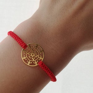 Op de afbeelding: Een rood koord armband met een gouden ronde bedel met een filigraan ontwerp.