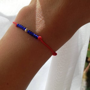 Pode incluir: Um bracelete de corda vermelha com contas de vidro azuis e uma conta prateada.