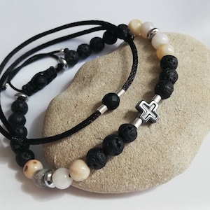 Może przedstawiać: Dwa bransoletki z czarnymi koralikami z lawy wulkanicznej, białymi i beżowymi koralikami oraz srebrnym wisiorkiem w kształcie krzyża. Jedna bransoletka ma czarny sznurek, a druga srebrny łańcuszek.