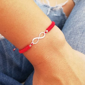 May include: Red string bracelet featuring a silver infinity symbol. The bracelet is made of a woven red cord with a silver infinity charm in the center. The bracelet is worn on a wrist.