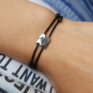 Puede incluir: Un brazalete de cuerda negra con dos dijes de flecha plateados. El brazalete es ajustable y tiene un diseño simple.