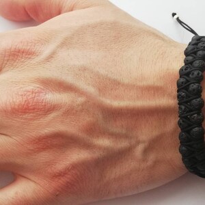 Puede incluir: Pulsera de piedra de lava negra con cierre de cord&oacute;n ajustable. La pulsera presenta dos filas de cuentas oscuras y texturizadas, conectadas por un cord&oacute;n negro. La pulsera se lleva en la mu&ntilde;eca, mostrando los tonos naturales y terrosos de la roca volc&aacute;nica.