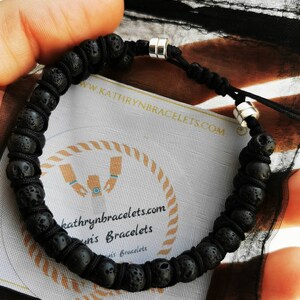 Puede incluir: Pulsera de piedra de lava negra con cord&oacute;n negro ajustable y detalles met&aacute;licos plateados. La pulsera se muestra en una tarjeta con el texto "kathrynbracelets.com" y "Kathryn's Bracelets". La pulsera se sostiene en una bolsa negra transparente.