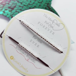 May include: Two black string bracelets with silver beads, one reads "For Him EVER" and the other reads "For Her FOREVER". The bracelets are displayed on a white card with the text "The Morse Code" and "Kathryn's Bracelets".