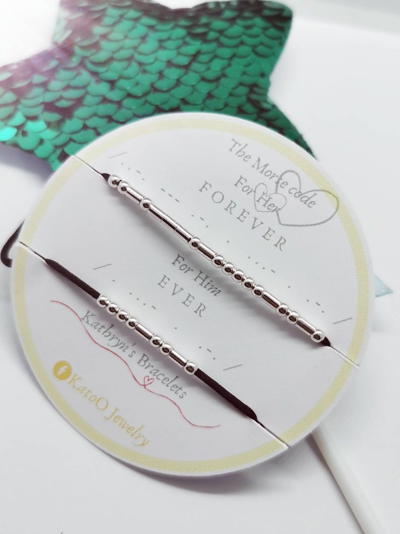 May include: Two black string bracelets with silver beads, one reads "For Him EVER" and the other reads "For Her FOREVER". The bracelets are displayed on a white card with the text "The Morse Code" and "Kathryn's Bracelets".