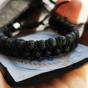 Puede incluir: Pulsera de piedra de lava negra con un dise&ntilde;o tejido. La pulsera est&aacute; hecha de cuentas negras redondas y porosas unidas con un cord&oacute;n negro. La pulsera se muestra sobre una superficie azul claro y blanca.