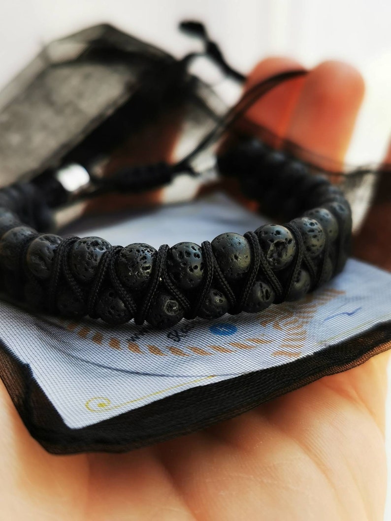 Puede incluir: Pulsera de piedra de lava negra con un dise&ntilde;o tejido. La pulsera est&aacute; hecha de cuentas negras redondas y porosas unidas con un cord&oacute;n negro. La pulsera se muestra sobre una superficie azul claro y blanca.