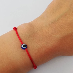 Può includere: Braccialetto di corda rossa con un ciondolo blu a forma di occhio turco. Il braccialetto è legato al polso di una persona.