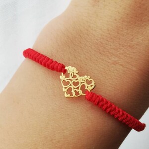 Puede incluir: Un brazalete de cuerda roja con un dije de corazón de oro. El dije de corazón está hecho de muchos corazones más pequeños.