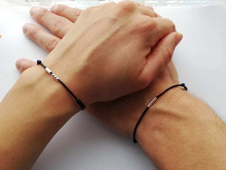 May include: Two black string bracelets with silver beads and a silver bar charm. The bracelets are worn on the wrists of two people.