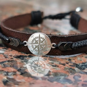 May include: A brown leather bracelet with a black cord and a silver compass charm. The compass has a black needle and a white face with black markings.