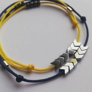 May include: A yellow and navy blue friendship bracelet with silver arrow-shaped beads.