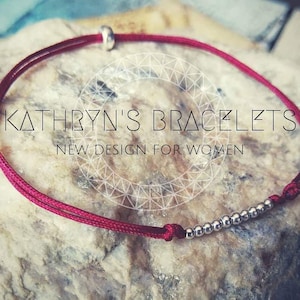 Puede incluir: Un brazalete de cuerda roja con cuentas plateadas. El brazalete está sobre una superficie de roca texturizada. El texto "KATHRYN'S BRACELETS NEW DESIGN FOR WOMEN" es visible en la imagen.