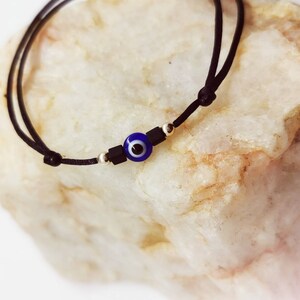 May include: A black cord bracelet with a blue evil eye charm and black beads. The bracelet is tied in a knot and is on a white rock.