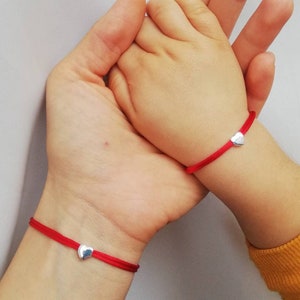 Puede incluir: Dos pulseras de cuerda roja con dijes de corazón plateados. Las pulseras se llevan en las muñecas de dos personas, una adulta y una niña, cuyas manos están entrelazadas.