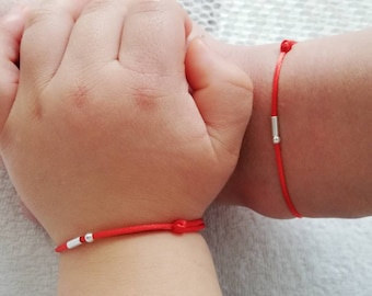 May include: Two red string bracelets with silver beads on a child's wrists.