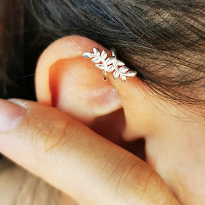 Könnte beinhalten: Ein silberner Ohrklemmen-Schmuck mit einem filigranen Blattdesign. Der Schmuck umschließt das Ohr und zeigt eine Ansammlung detaillierter Blätter. Der Schmuck wird am Ohr getragen, mit Haut und Haaren im Hintergrund.