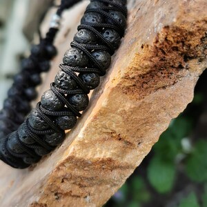 Puede incluir: Pulsera de roca volc&aacute;nica negra con un dise&ntilde;o de cord&oacute;n trenzado. La pulsera presenta cuentas de lava redondas y porosas ensartadas con un cord&oacute;n negro. La pulsera se muestra sobre una superficie de piedra marr&oacute;n claro.