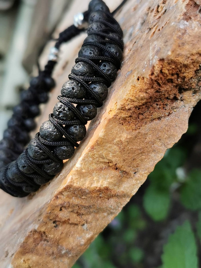 Puede incluir: Pulsera de roca volc&aacute;nica negra con un dise&ntilde;o de cord&oacute;n trenzado. La pulsera presenta cuentas de lava redondas y porosas ensartadas con un cord&oacute;n negro. La pulsera se muestra sobre una superficie de piedra marr&oacute;n claro.