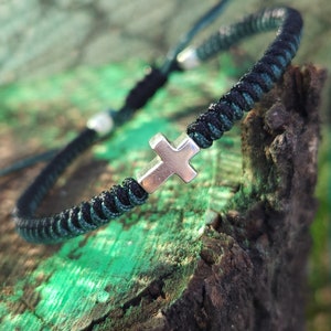 May include: A black and green braided bracelet with a silver cross charm. The bracelet is tied with a knot and has a silver bead on the end.