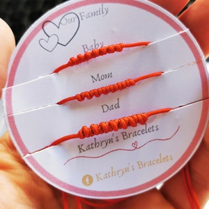 Könnte beinhalten: Eine weiße Karte mit den Worten "Our Family" und "Kathryn's Bracelets" darauf gedruckt. Auf der Karte befinden sich vier rote Armbänder aus Schnur, beschriftet mit "Baby", "Mom", "Dad" und "Kathryn's Bracelets".