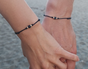 Puede incluir: Dos personas se toman de la mano, cada una lleva una pulsera de cuerda negra con una cuenta dorada y una cuenta cuadrada negra.