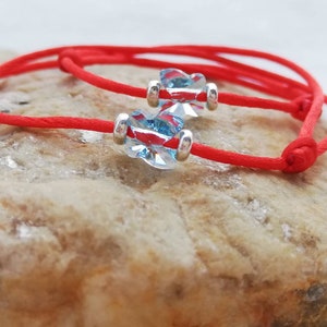 May include: Two red string bracelets with light blue crystal beads. The bracelets are tied together and are lying on a light brown rock.