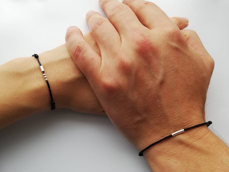 May include: Two black string bracelets with silver beads and a silver bar charm. The bracelets are worn on the wrists of two people.